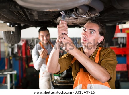 Mechanics in orange and white coveralls are repairing exhaust system of a car. Workmen replacing a muffler on a sedan