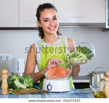 Happy smiling adult girl steaming salmon and vegetables in domestic kitchen