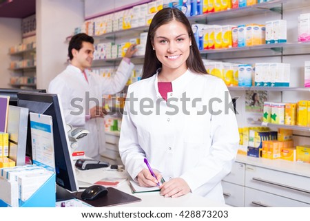 Cheerful pharmacist standing at pay desk and pharmacy technician helping