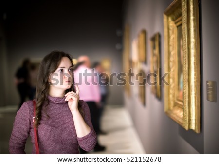 Portrait of positive young girl attentively looking at paintings in art museum