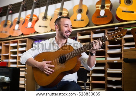 Joyful smiling young guy choosing acoustic guitar in music instruments shop