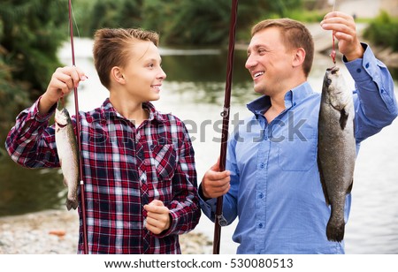 Portrait of glad teenage boy and his father holding fish on hook in hands