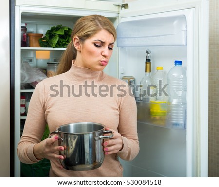 Upset blonde woman holding pot with foul meal near fridge