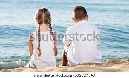 portrait of boy and girl sitting back forward together on sandy beach in sunlight