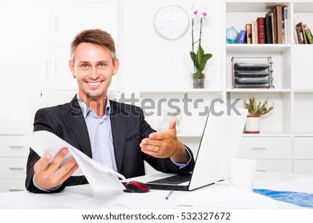 Smiling professional man sitting with laptop and having paper document in hands at firm office