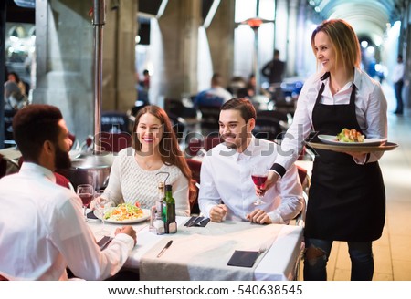 Portrait of smiling adult friends in outdoors restaurant and smiling waitress.  Focus on blonde girl