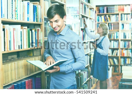 Young smiling cheerful  man reading book while choosing it in book shop
