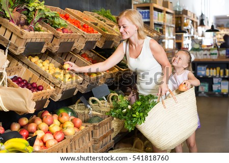 happy smiling female customer with little daughter choosing fruits in food shop