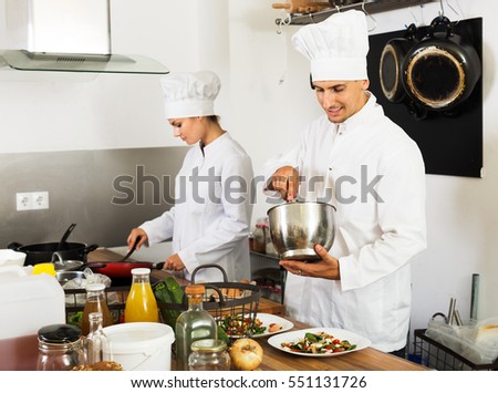 Glad young man and woman in cook uniform preparing food at kitchen and smiling