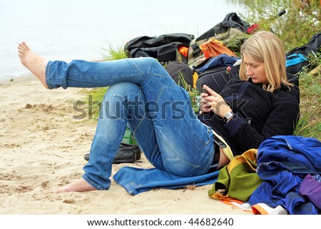 bored barefoot young blond women gaming on mobile phone, shore, sand, grass and bags on background