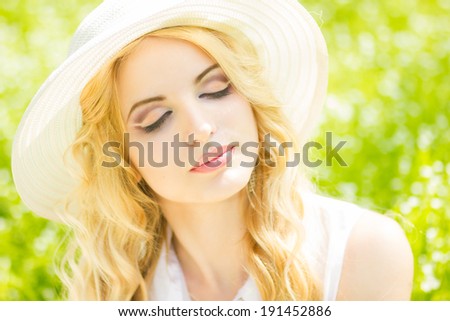 Portrait of a beautiful young blonde woman with wavy hair in nature. Girl in white hat sitting on the grass in the park