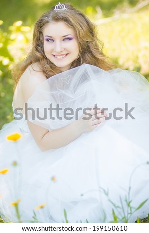 Portrait of beautiful young bride with long curly hair. Young girl sitting in a wedding dress in the park among yellow flowers.