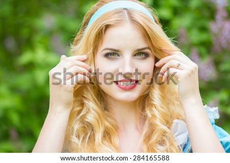 Portrait of a beautiful young blonde woman with long hair dressed as Alice in Wonderland. Girl on the nature near the lilac bushes. Soft focus
