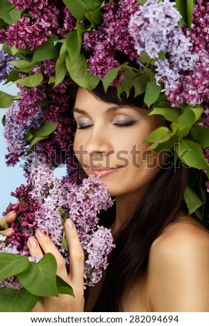 Beautiful young model woman with lilac bouquet and wreath as flowers hair style sniffs the sweet smell of aroma on blue background