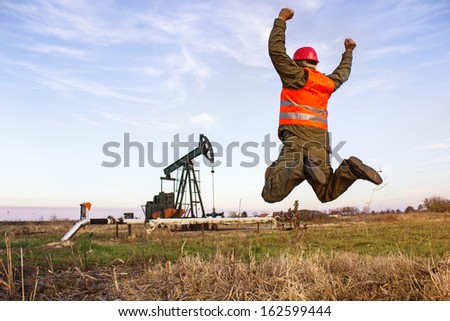 Oil Industry Pump jack with one oil worker jumping with joy,best focus on prslku, pockets of pants right hand man and the grass below, copi space, blurred shoes