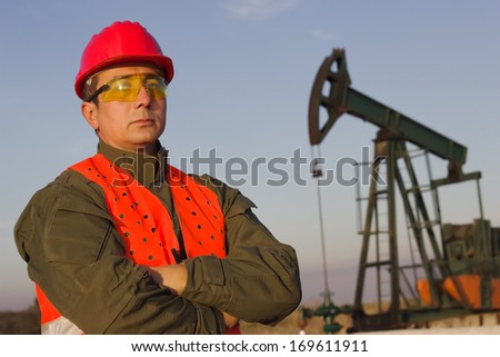 worker on an oil pump stands with arms crossed and looking at the camera, best focus on the man, background blurred
