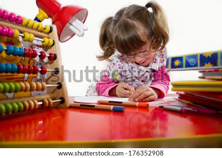 Cute little girl with glasses is drawing with felt-tip pen in preschool,best focus glasses, hair, shirt and pencil in hand, soft focus device for computing the pen on the table, books, hand