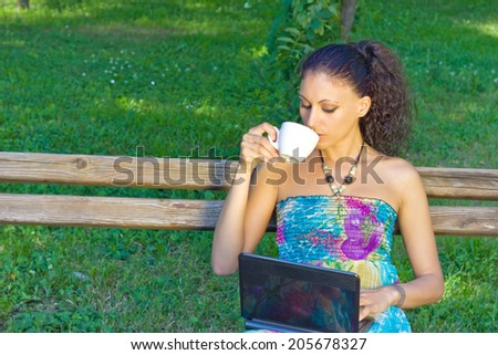 beautiful girl with black hair sitting on a bench drinking coffee and looking at laptop, best focus on the girl, soft focus background