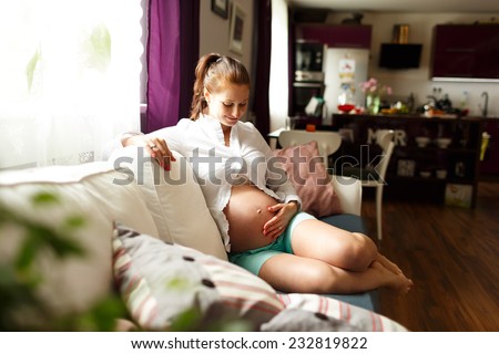 Pregnant woman sitting relax on bed. Sitting on sofa. Pregnant woman touching her belly with hands, relaxing at home on the couch, pregnant mother keeping her hands on belly while looking at it