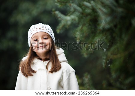 Adorable little girl posing near the branch of tree and smiling in to a camera. Wearing winter coat and hat. Lovely young girl in the winter outdoors.