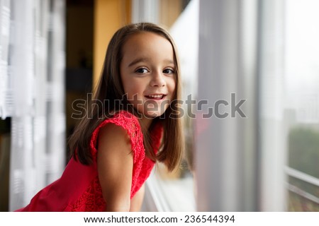 Beautiful little girl smiling and watching out the window. A child looks out the window. Young girl looking from window. Portrait of cheerful kid sits at window.