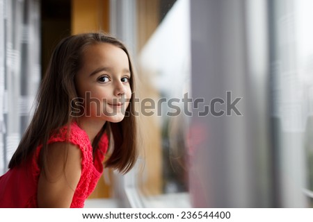 Beautiful little girl smiling and watching out the window. A child looks out the window. Young girl looking from window. Portrait of cheerful kid sits at window.