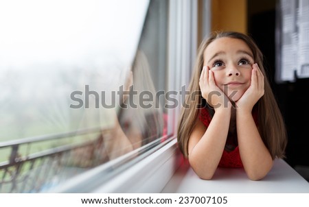 Beautiful little girl smiling and watching out the window. A child looks out the window. Young girl looking from window. Portrait of cheerful kid lies at windowsill.