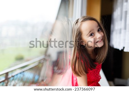 Beautiful little girl smiling and watching out the window. A child looks out the window. Young girl looking from window. Portrait of cheerful kid sits at windowsill.