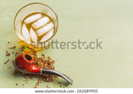Tobacco pipe on rustic warn green wood surface with spilled natural tobacco and a glass of whisky on the rocks