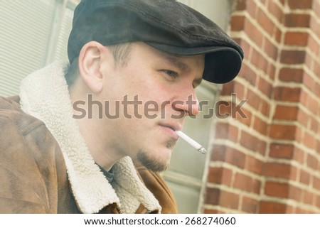 Cool guy in aviator jacket and newsie cap relaxes against a glass wall and enjoys his cigarette