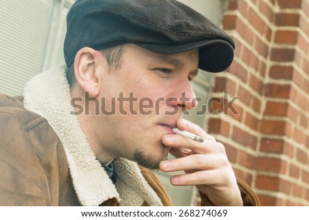 Cool guy in aviator jacket and newsie cap relaxes against a glass wall and enjoys his cigarette
