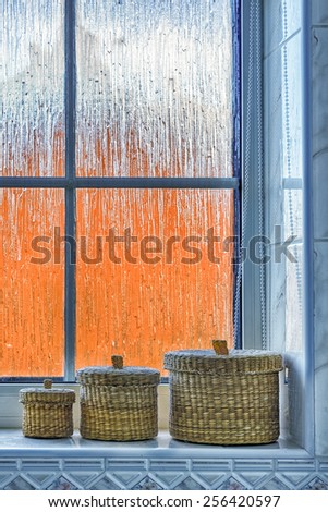Set of three small wicker baskets sitting on a bathroom windowsill against a frosted window, with distorted orange exterior [portrait format].