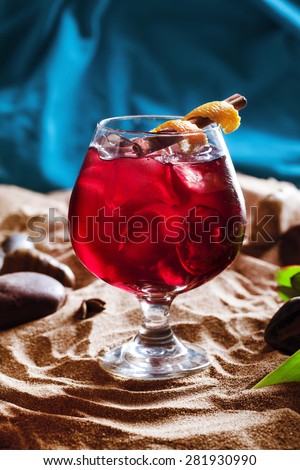 red cocktail with ice cinnamon and orange peel, masculine style in the sand on a blue background