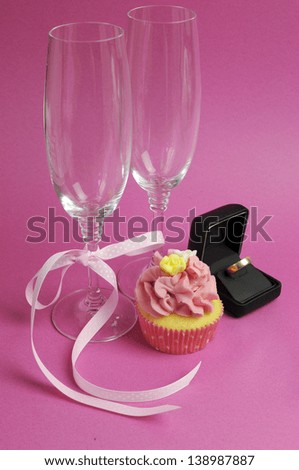 Wedding theme bridal pair of champagne flute glasses with pink cupcake and wedding ring in black jewelry box against a pink background. Vertical.