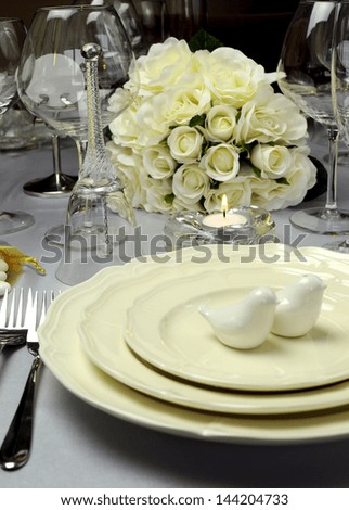 Close up of detail on wedding breakfast dining table setting with dove shape salt and pepper shakers on beautiful china plates with white roses bouquet in background. Vertical.