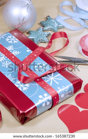 Christmas gift wrapping with red, white and aqua blue wrapping, hearts, tags, scissors and gift boxes with festive Christmastime ornament decorations on a pale wood table.  Selective focus. Vertical.