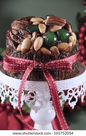 Festive Christmas food, fruit cake with glace cherries and nuts on white cake stand with holiday berry wreath in green background. Selective focus. Close up vertical.