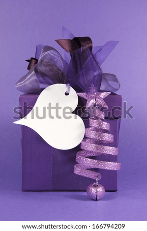 Festive purple gift box present with heart shape gift tag and Christmas bell decoration ornament, on purple background.