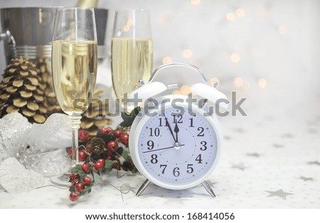 Happy New Year table setting with white retro clock showing five to midnight with champagne and festive decorations against white starry background with bokeh lights.