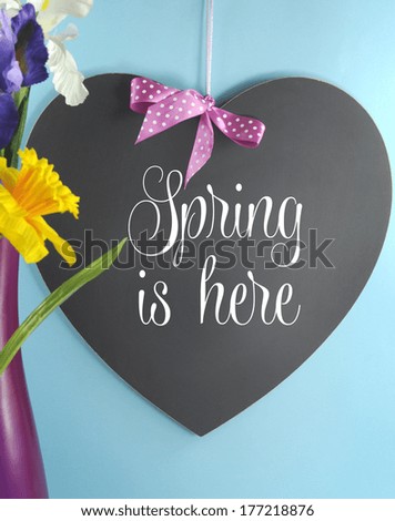 Spring Is Here greeting on heart shape blackboard or copy space with spring flowers in pink vase on blue background. Vertical.