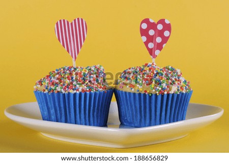 Bright and cheery red blue and yellow theme cupcakes with hundred and thousands candy topping and heart toppers for birthday or special occasion on yellow background.