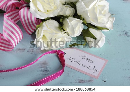 Happy Mothers Day gift of white roses bouquet with pink stripe ribbon and gift tag with greeting on aqua blue vintage shabby chic table.