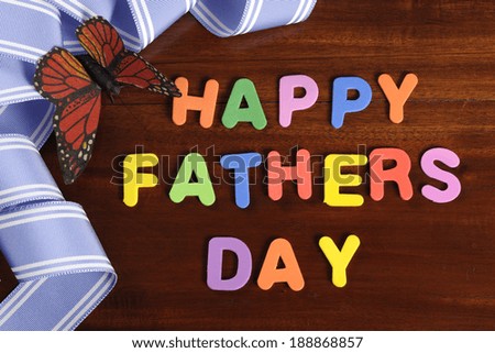 Happy Fathers Day childrens toy block colorful letters spelling greeting on dark rustic wood table with blue ribbon and butterfly.