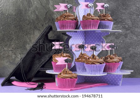 Graduation day pink and purple party cupcakes, with chocolate frosting, on polka dot three tier stand and large graduation cap against modern black slate kitchen tiles.