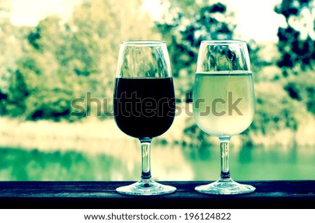 Retro filter style two glasses of wine, white and red, on wooden rail with country rural scene in background.
