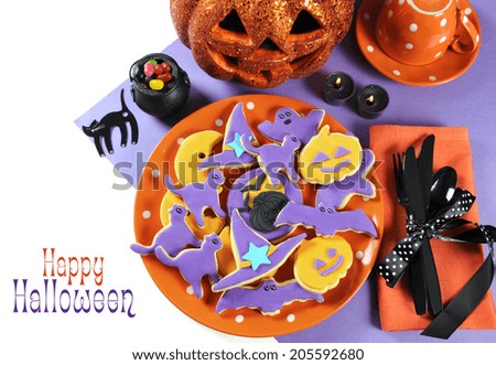 Happy Halloween orange and purple sugar cookies in cat, hat, bat and pumpkin shapes on orange polka dot plate as party table centerpiece with jack o lantern pumpkin, black cat and sample text.