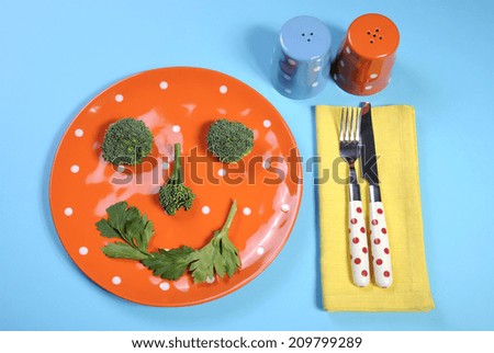 Healthy diet health food concept with happy smiley face made from broccoli and celery on a fun orange polka dot plate with salt and pepper shakers, and colorful cutlery on a pale blue background.