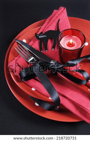 Elegant red and black theme Halloween party dining Table place setting with plates, cutlery, black cat and lit candle. Vertical.