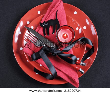 Elegant red and black theme Halloween party dining Table place setting with plates, cutlery, black cat and lit candle.