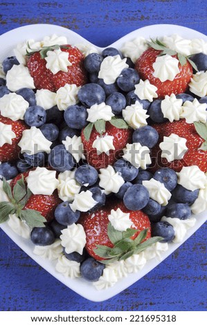 Patriotic red, white and blue berries with fresh whipped cream stars in white heart shape flag on rustic dark blue wood background. Vertical elevated aerial view.